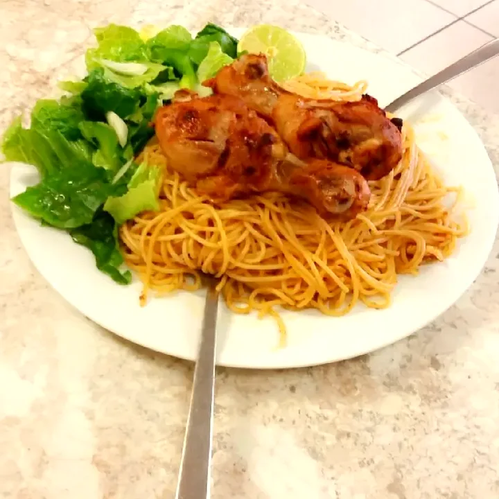 A plate of spaghetti topped with roasted chicken legs, with green salad and a lime wedge